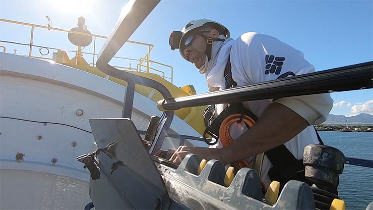 A person leaning over a wave energy converter on a boat in the sun.