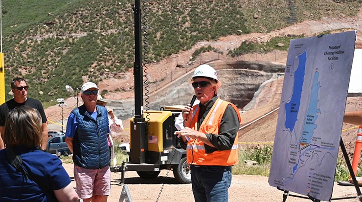 A person smiles while wearing safety glasses and a hard hat in front of a body of water, a forest, and a mountain
