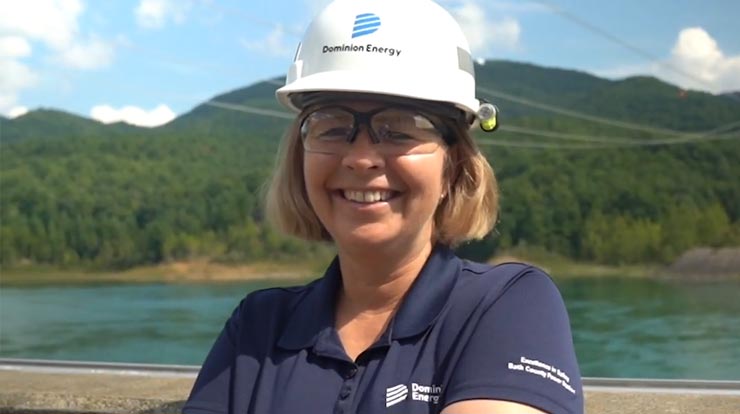 A person smiles while wearing safety glasses and a hard hat in front of a body of water, a forest, and a mountain