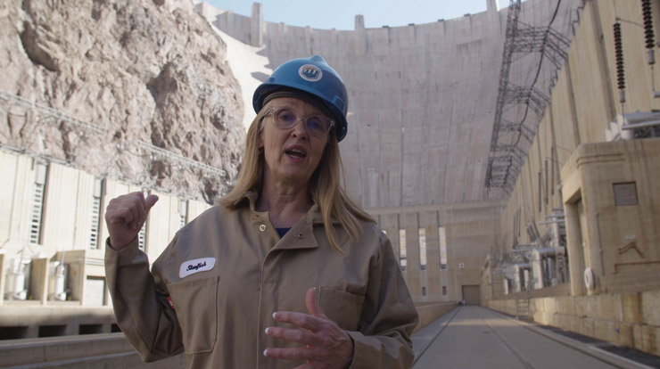 A person wearing a hard hat and safety glasses stands in front of a tall dam and points