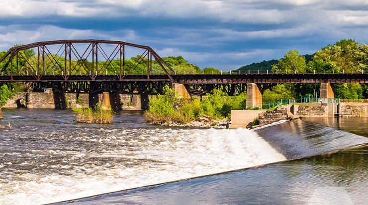 A shallow dam and lake with an elevated train track in the background with blue sky and trees 