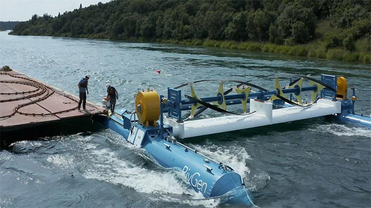 A long hydrokinetic device sits on the riverbed, with two people working on an adjacent platform.