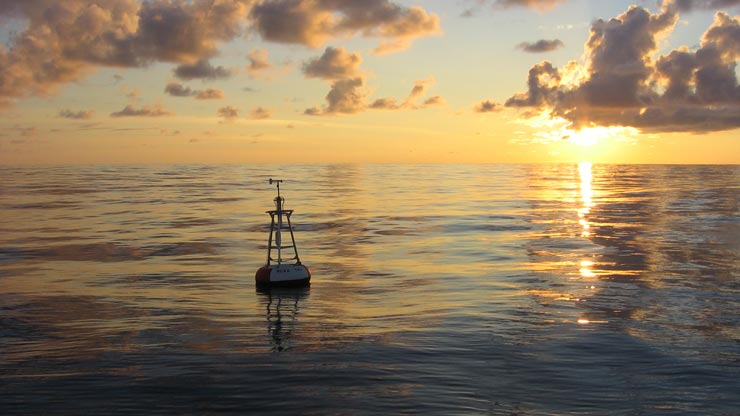 A buoy in the ocean with the sun setting in the background and a blue-yellow sky filled with clouds