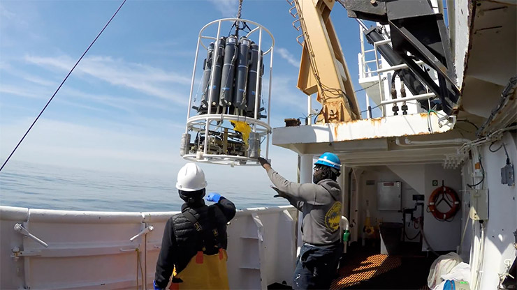 Two people on a vessel with a black device hanging on cables over them