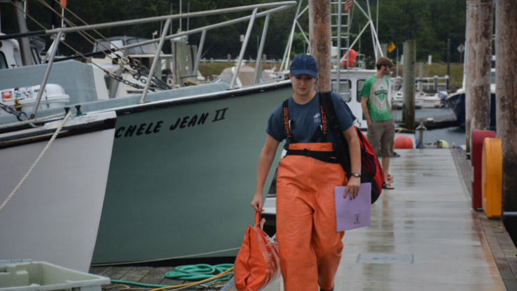 A person in orange work gear walking toward the camera on a dock with boats in the background