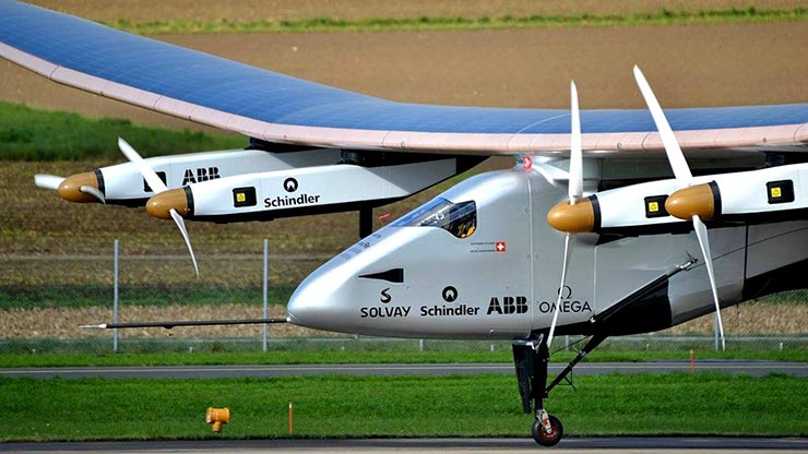 A silver, two-propeller plane with black logos in front of farmland.