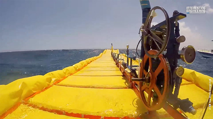 A long, yellow marine energy device with mechanical wheels floating on the ocean surface.