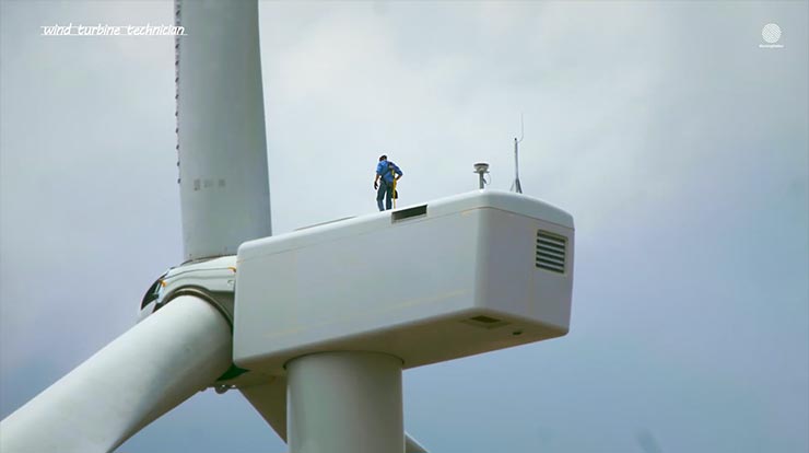 A person standing on top of a large wind turbine with a grayish-blue sky in the background.